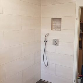 Barrierefreie, moderne ebenerdige Dusche mit großen hellen Fliesen und Nischenablage.