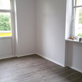 Helles, leeres Zimmer mit weißen Wänden, Laminatboden und zwei großen Fenstern.